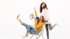 two-happy-girls-in-sweaters-having-fun-with-shopping-trolley-and-megaphone-over-white-wall