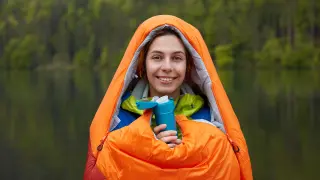 Smiling female traveler being in good mood, wrapped in sleeping bag, spends free time at nature, holds thermos of hot drink, warms near river during cold day. People, receation, lifestyle concept