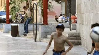 Un joven sin camiseta patina junto a las terrazas de la plaza de San Bruno, mientras otros juegan al fútbol.