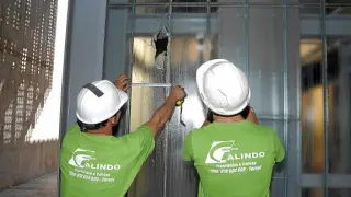Dos operarios toman medidas para reponer el cristal de la caja del ascensor roto de un botellazo.