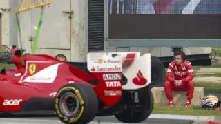 - El piloto español Fernando Alonso, de la escudería Ferrari, observa sentado fuera de pista cómo una grúa retira su monoplaza