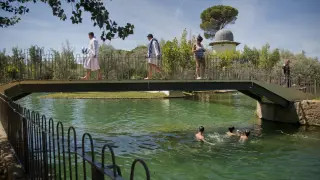Unos bañistas pasan sobre el puente del lago de un balneario de Alhama de Aragón.