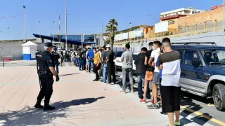 Varios migrantes procedentes de Marruecos hacen cola en las puertas de la frontera de Ceuta donde agentes de la Policía Nacional reparten citas para solicitud de asilo.