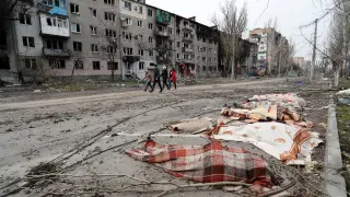 Cuatro civiles caminan por una calle de Mariúpol donde se ven varios cadáveres