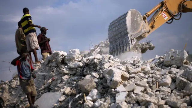 Unos haitianos observan una grúa desde una montaña de escombros, un año después de la tragedia