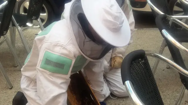 Dos bomberos colocan una caja con una panal de cera junto a un enjambre de abejas que estaba en la silla de un velador