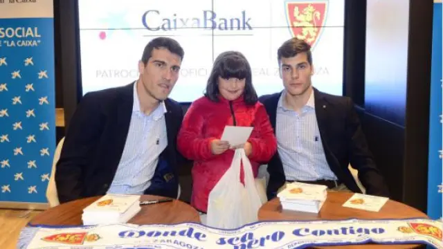 Zapater y Soro, junto a una niña, en el estreno de la campaña "Ningún niño sin juguete", este martes en la sede de Caixa Bank Store en la Plaza Aragón.