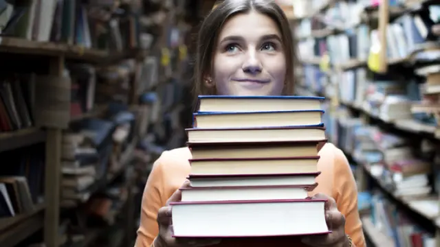 young girl student holds a stack of books in the library, she searches for literature and offers to read, a woman prepares for study, bookseller on the background of the bookstore, knowledge is power, concept