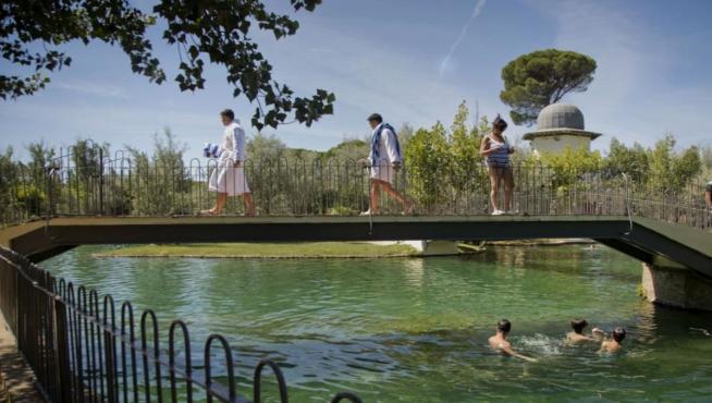 Unos bañistas pasan sobre el puente del lago de un balneario de Alhama de Aragón