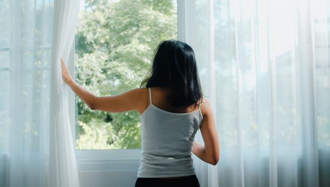 Happy beautiful Asian woman smiling and drinking a cup of coffee or tea near the window in bedroom. Young latin girl open curtains and relax in morning. Lifestyle lady at home concept.