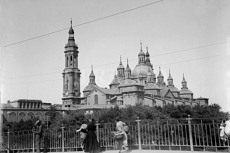 Vista del templo del Pilar de Zaragoza (fue Basílica en 1948), con solo dos torres, desde el puente de Piedra en 1929. En la fotografía, se ve como las mujeres cruzaban el puente, cargadas con sus cestas, para ir a atender sus faenas. La barandilla metálica colocada en 1908 pervivió en el puente hasta 1991