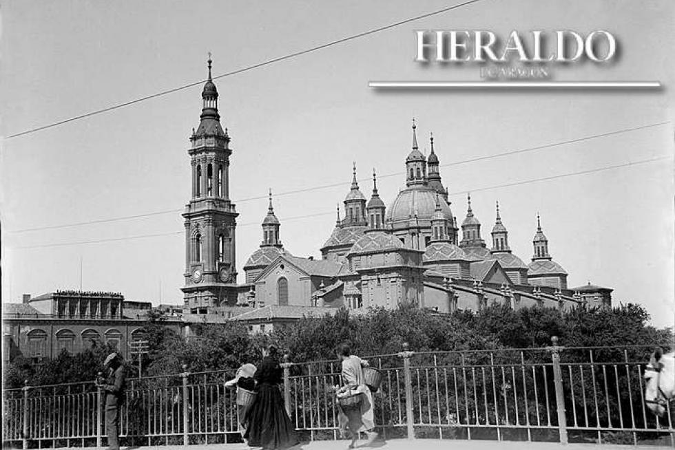 Vista del templo del Pilar de Zaragoza, con solo dos torres, desde el puente de Piedra en 1929. En la fotografía, se ve como las mujeres cruzaban el puente, cargadas con sus cestas, para ir a atender sus faenas. La barandilla metálica colocada en 1908 pervivió en el puente hasta 1991. Fotografía incluida en el coleccionable 'Cómo hemos cambiado' que HERALDO DE ARAGÓN regala en los meses de octubre y noviembre a sus lectores.