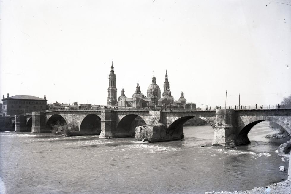 Peatones cruzan el puente de piedra con la Basílica del Pilar al fondo