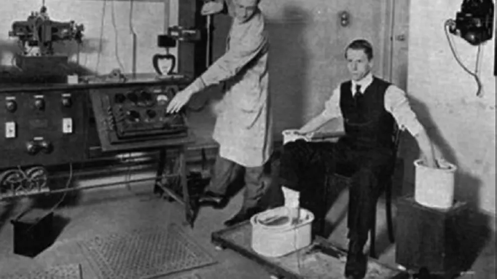 Willem Einthoven prueba el galvanómetro de hilo con un paciente, con manos y pies desnudos dentro de cubetas de disolución salina (1916)