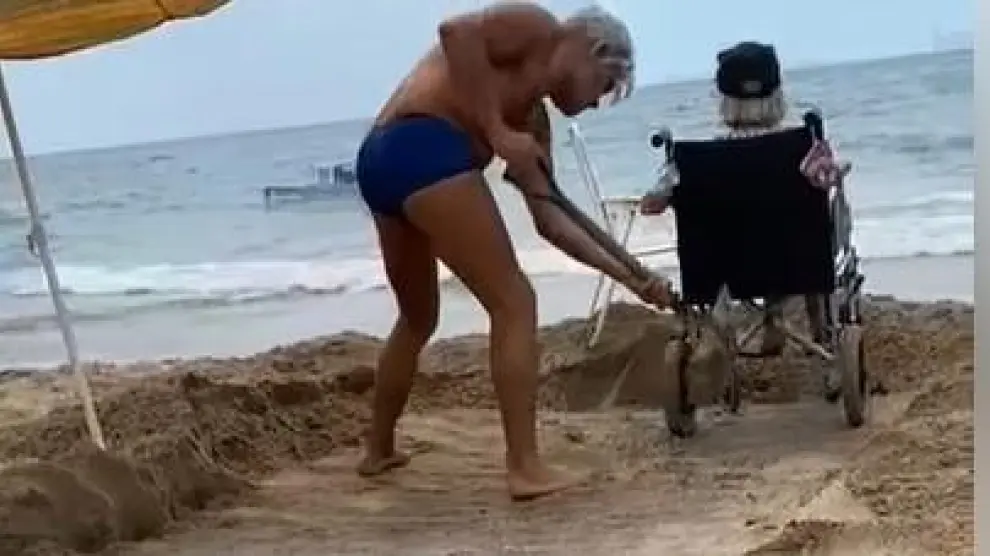 El hombre limpia con una pala la pasarela, cubierta de arena, para que su madre pueda acceder al mar.