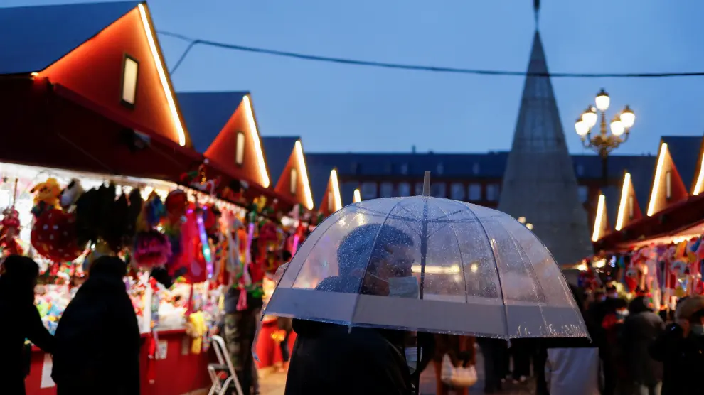 Un joven con mascarilla camina bajo un paraguas en el mercado navideño de la plaza Mayor de Madrid