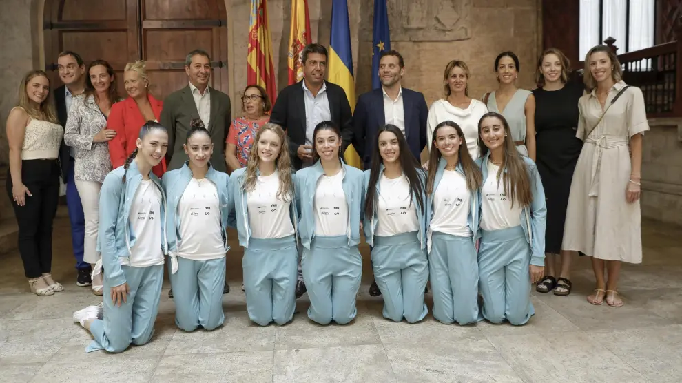 Fotografía del Gobierno valenciano junto a las integrantes de la selección de gimnasia rítmica en el que las deportistas aparecen posando de rodillas en fila delante de los políticos.