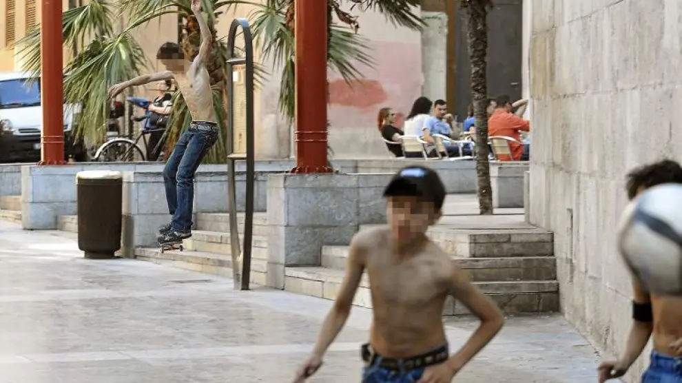 Un joven sin camiseta patina junto a las terrazas de la plaza de San Bruno, mientras otros juegan al fútbol.
