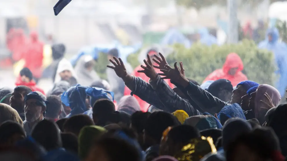 Un grupo de refugiados alza la mano para intentar alcanzar un chubasquero que les aísle de la lluvia.