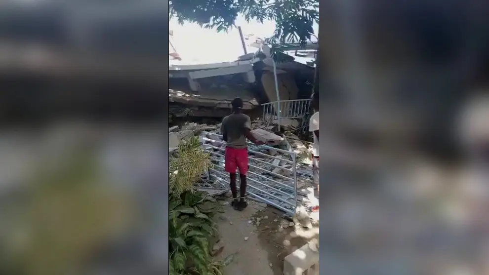 People stand in front of a collapsed building following an earthquake, in Les Cayes, Haiti, in this still image taken from a video obtained by Reuters on August 14, 2021.   REUTERS TV via REUTERS THIS IMAGE HAS BEEN SUPPLIED BY A THIRD PARTY. MANDATORY CREDIT.[[[REUTERS VOCENTO]]] HAITI-QUAKE/