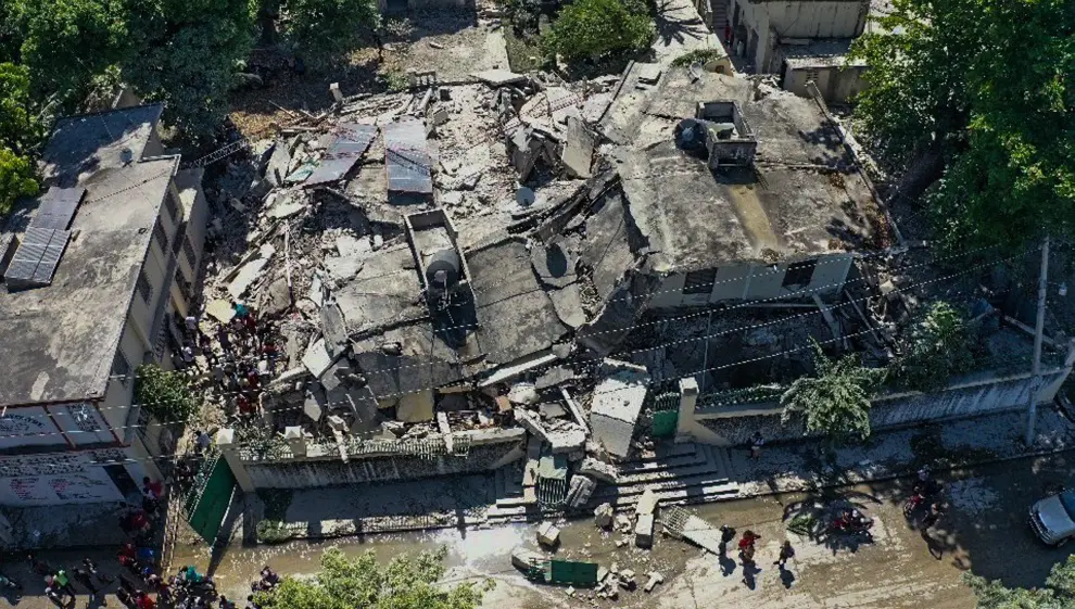 People stand in front of a collapsed building following an earthquake, in Les Cayes, Haiti, in this still image taken from a video obtained by Reuters on August 14, 2021.   REUTERS TV via REUTERS THIS IMAGE HAS BEEN SUPPLIED BY A THIRD PARTY. MANDATORY CREDIT.[[[REUTERS VOCENTO]]] HAITI-QUAKE/