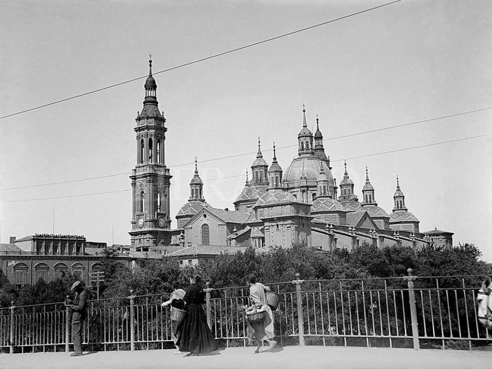 Vista del templo del Pilar de Zaragoza (fue Basílica en 1948), con solo dos torres, desde el puente de Piedra en 1929. En la fotografía, se ve como las mujeres cruzaban el puente, cargadas con sus cestas, para ir a atender sus faenas. La barandilla metálica colocada en 1908 pervivió en el puente hasta 1991