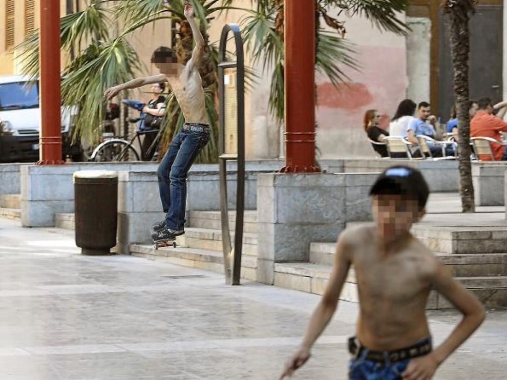 Un joven sin camiseta patina junto a las terrazas de la plaza de San Bruno, mientras otros juegan al fútbol.