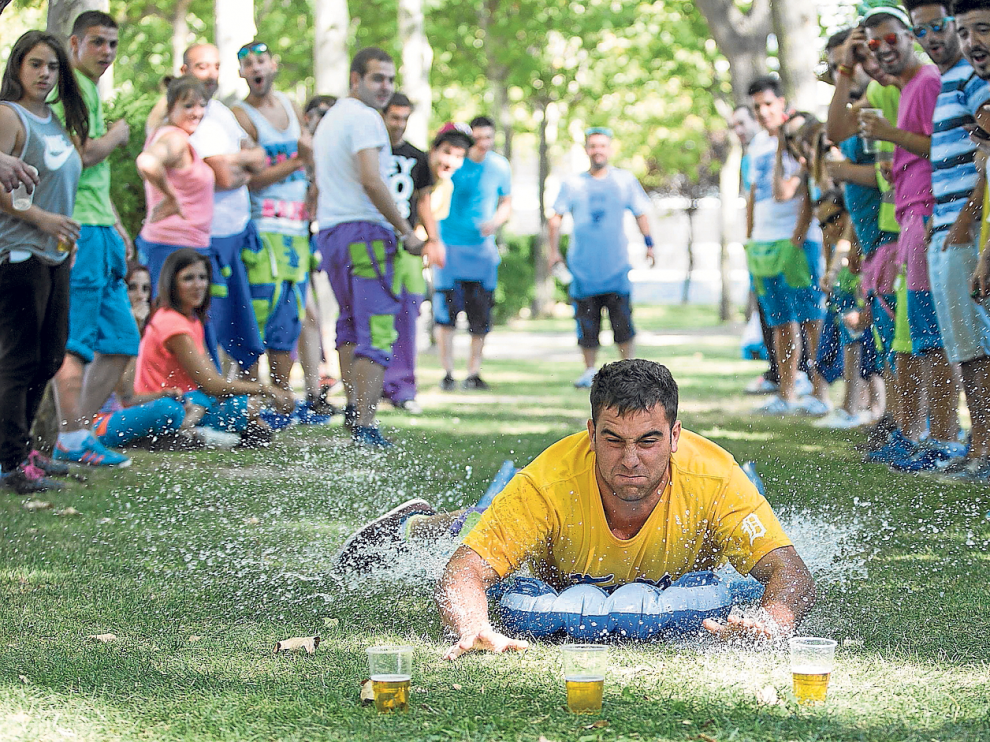 Un peñista se desliza con un flotador por una superficie de plástico llena de agua mientras intenta alcanzar un vaso de cerveza.
