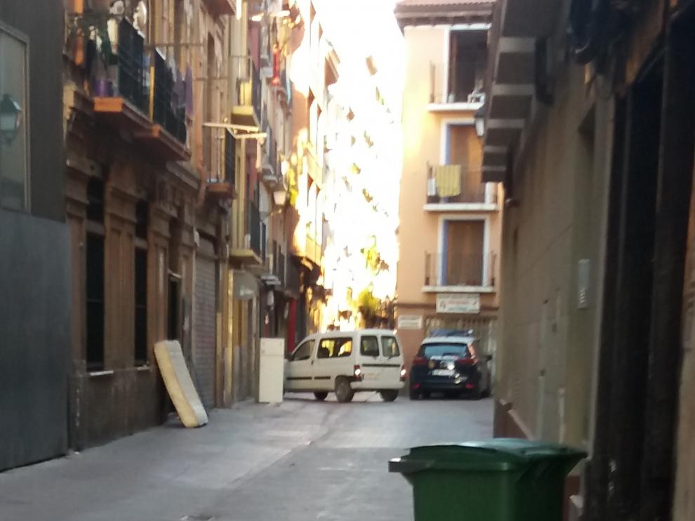 En primer plano, un contenedor rodeado de basura, al fondo, un colchón y un mueble abandonados.