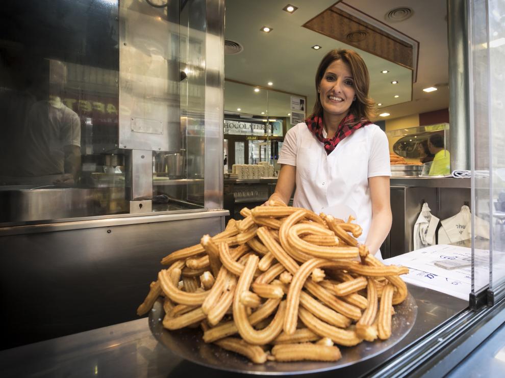 Raquel, ataviada con el cachirulo propio de estas fechas, sonríe tras una de las inmensas montañas de churros que caracterizan a La Fama. La imagen está tomada desde la calle, a través del ventanal que emplean para despachar docenas y docenas del manjar que prepara la familia desde 1949.