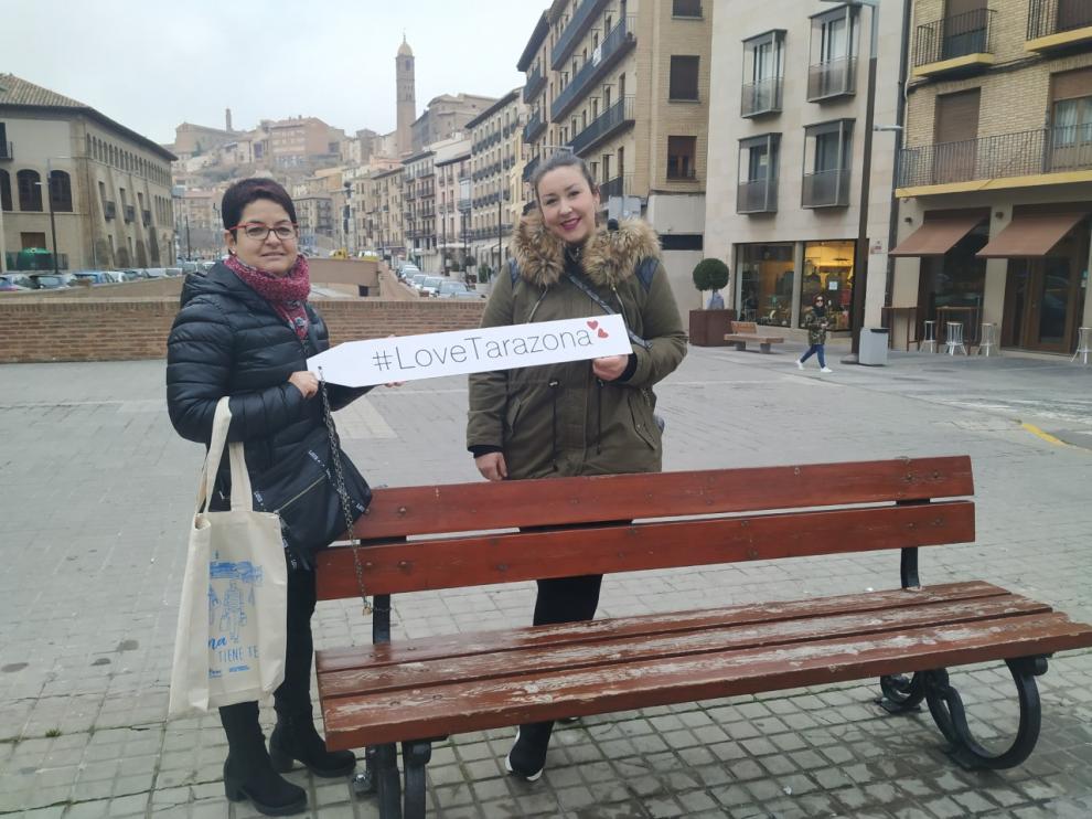 La gerente y la presidenta de ACT posan con el cartel de la campaña en uno de los bancos del concurso