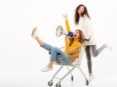 two-happy-girls-in-sweaters-having-fun-with-shopping-trolley-and-megaphone-over-white-wall